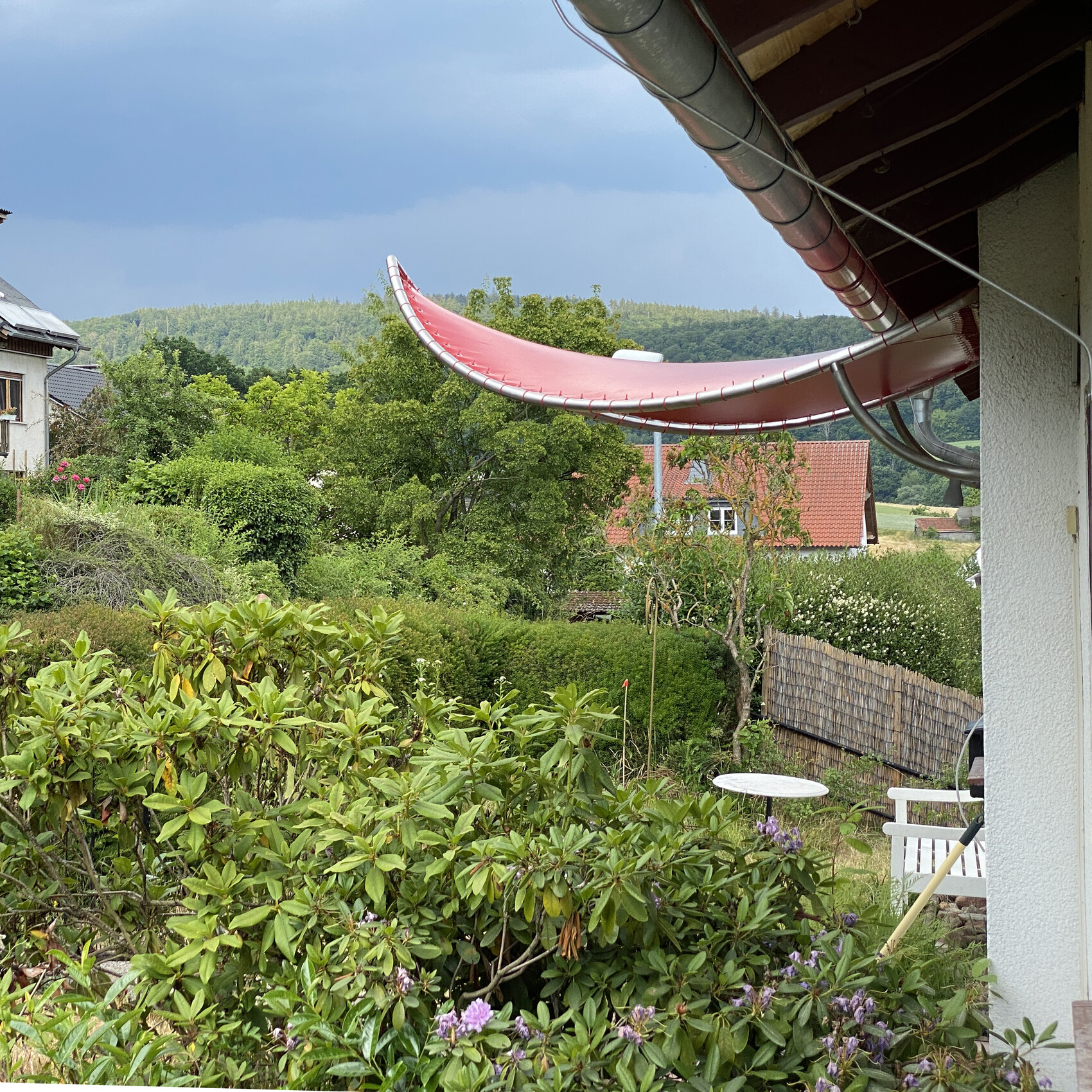 Garten mit dichtem Grün, roter gebogener Überhang am Haus und Hügel im Hintergrund