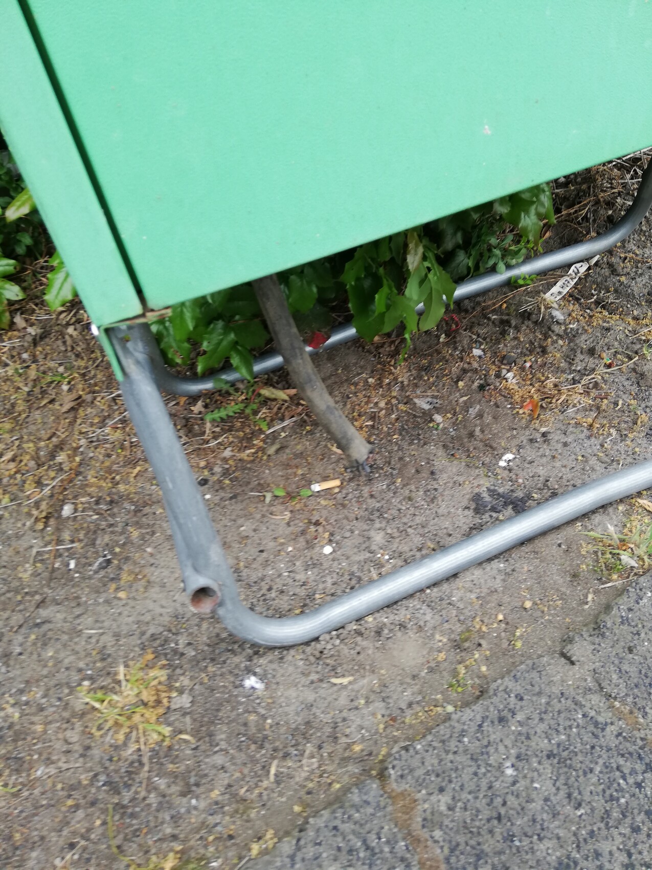 Grüner Metallkasten auf Beinen, daneben gebogenes Rohr; Boden mit Erde und Pflanzen.
