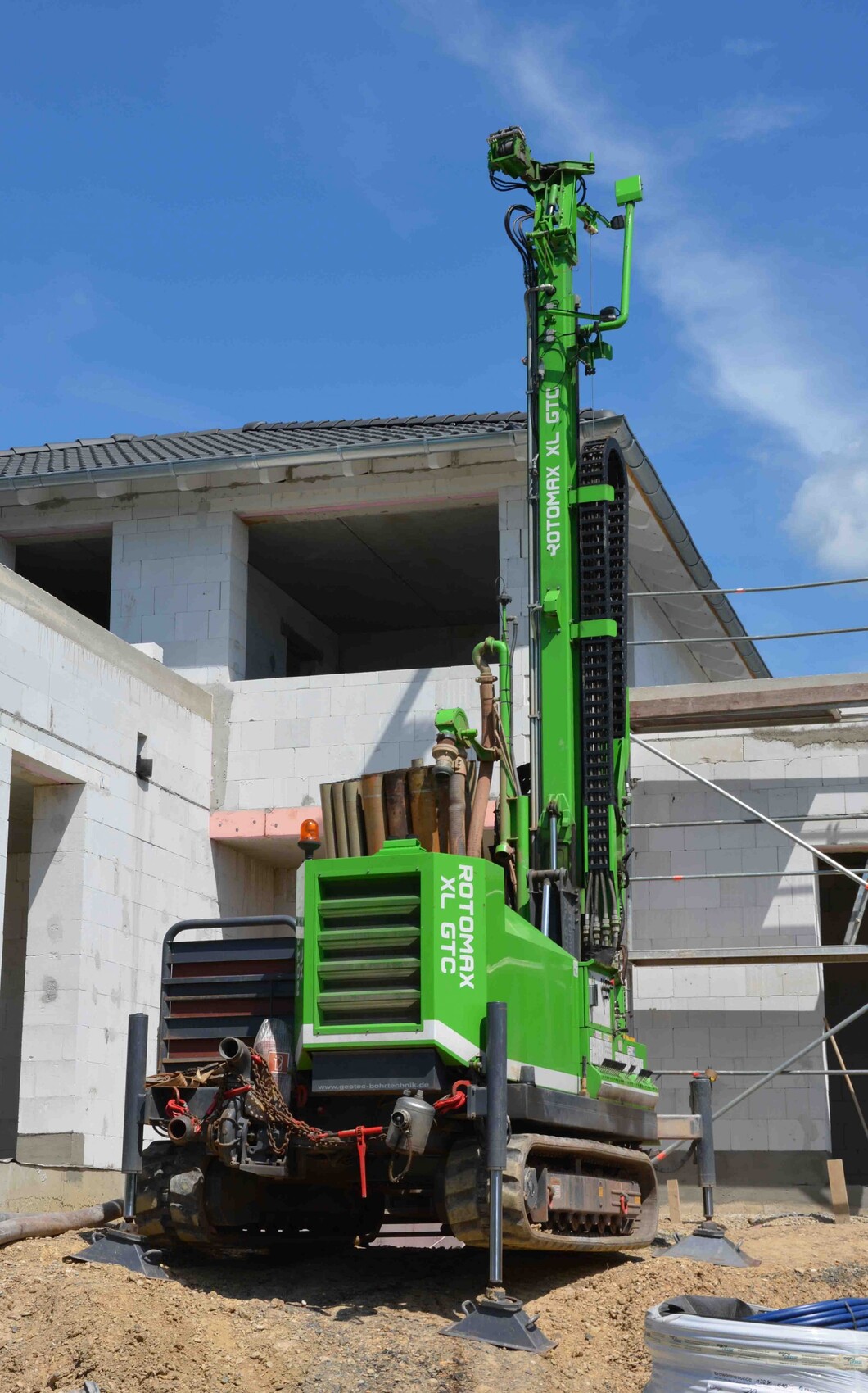 Grüne Bohranlage mit Ausleger auf Baustelle neben unfertigem Gebäude, blauer Himmel.