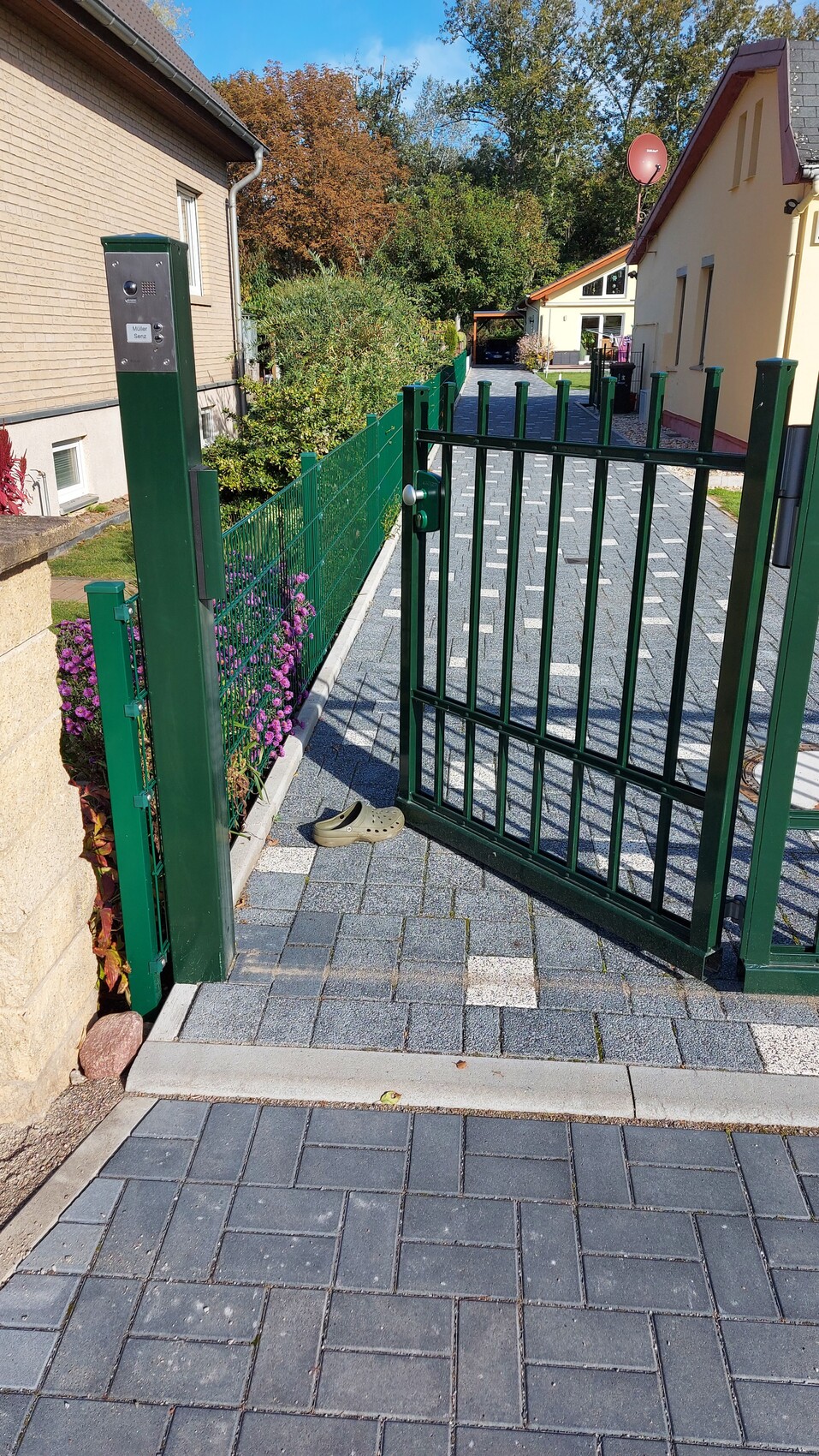 Grünes Einfahrtstor am gepflasterten Gartenweg mit Zaun und Häusern im Hintergrund