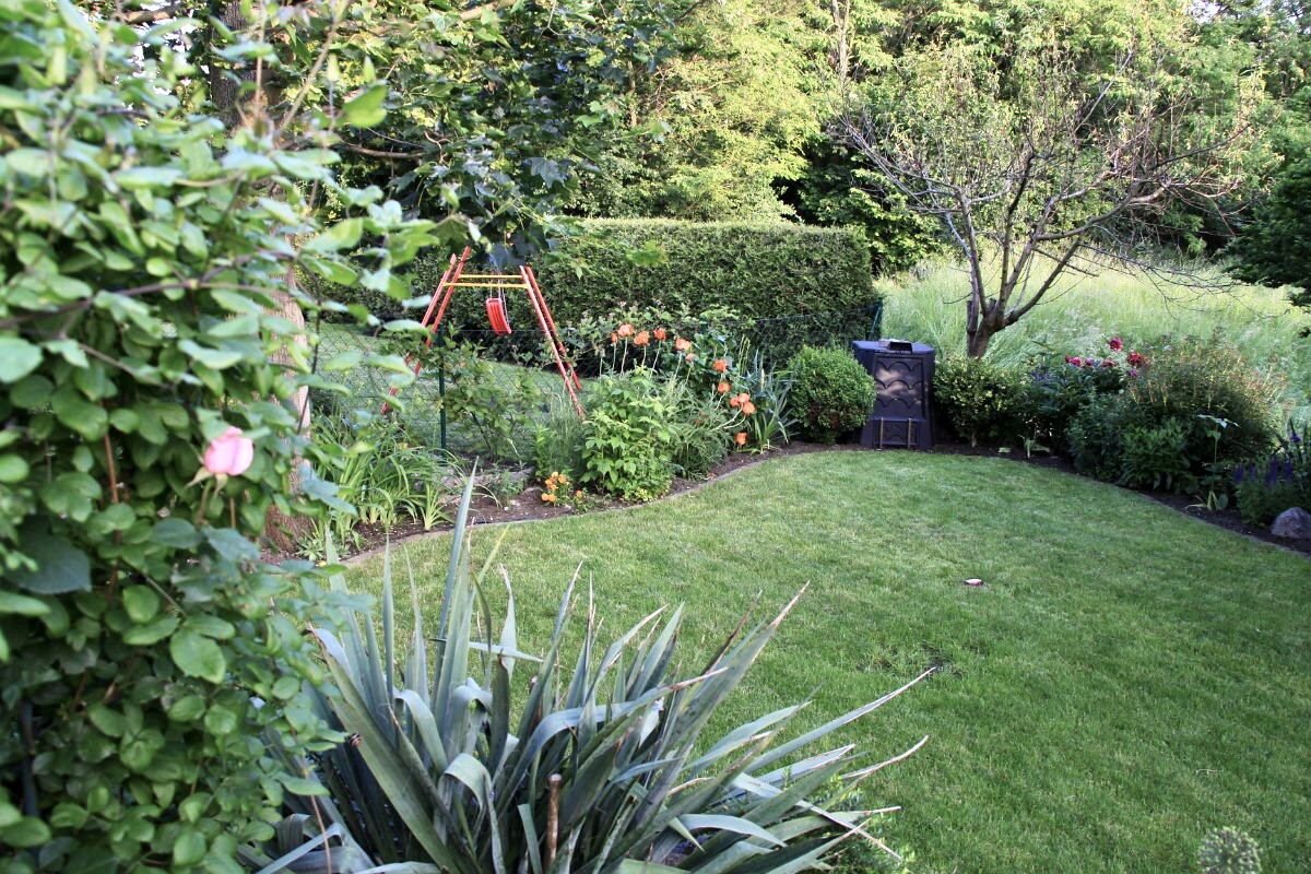Garten mit grünem Rasen, Hecken, Agaven, Rosen und rotem Schaukelrahmen hinter Blumenrabatten.