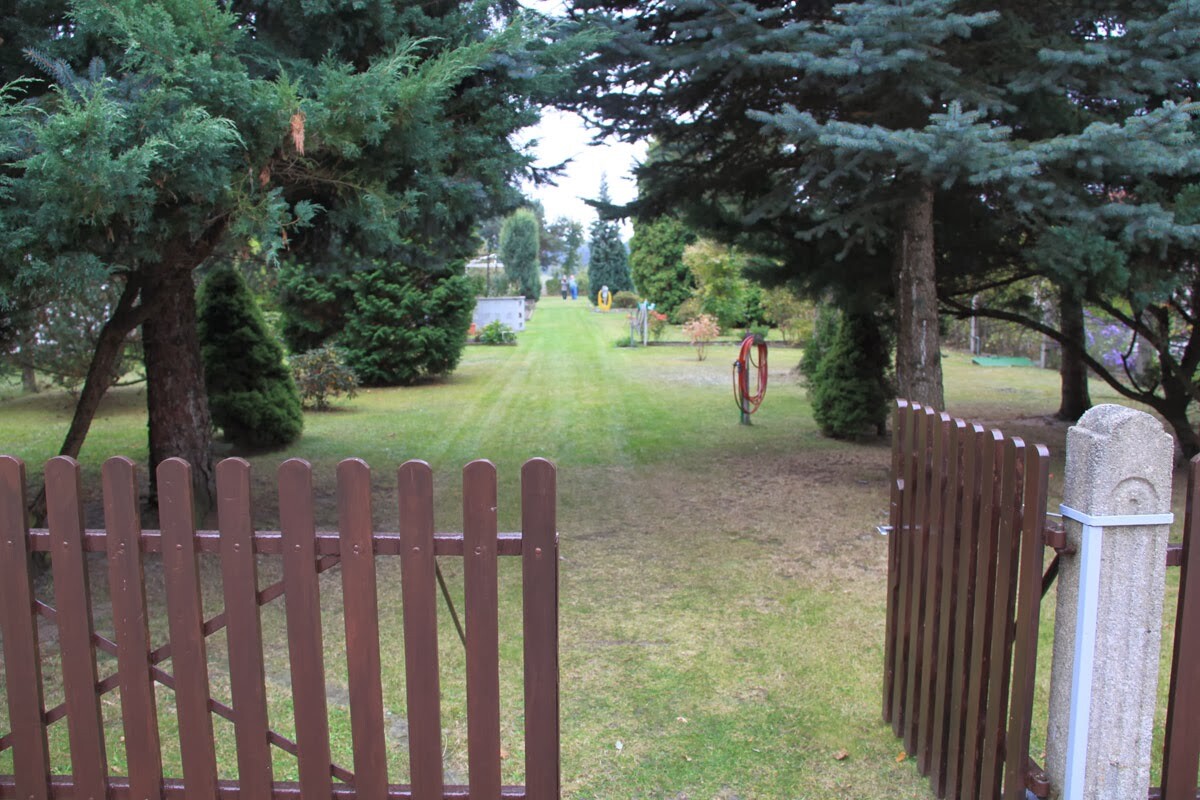 Gartenweg durch Park mit gemähter Wiese, brauner Holzzaun im Vordergrund, roter Schlauchhalter rechts