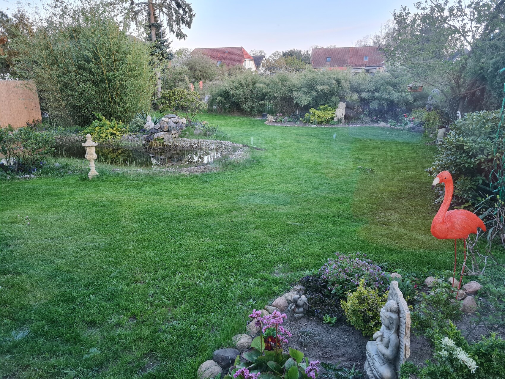 Gruener Garten mit Rasen, Skulpturen und rotem Flamingo-Deko im Vordergrund