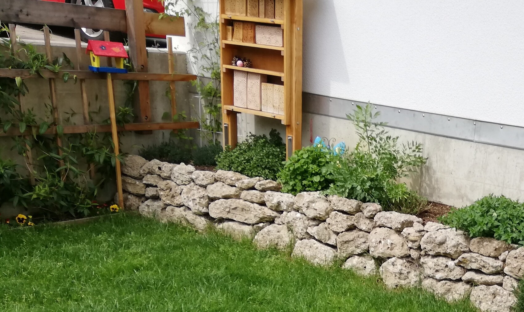 Garten mit Gras, Steinmauer aus Natursteinen, Holzregal und Vogelhaus.