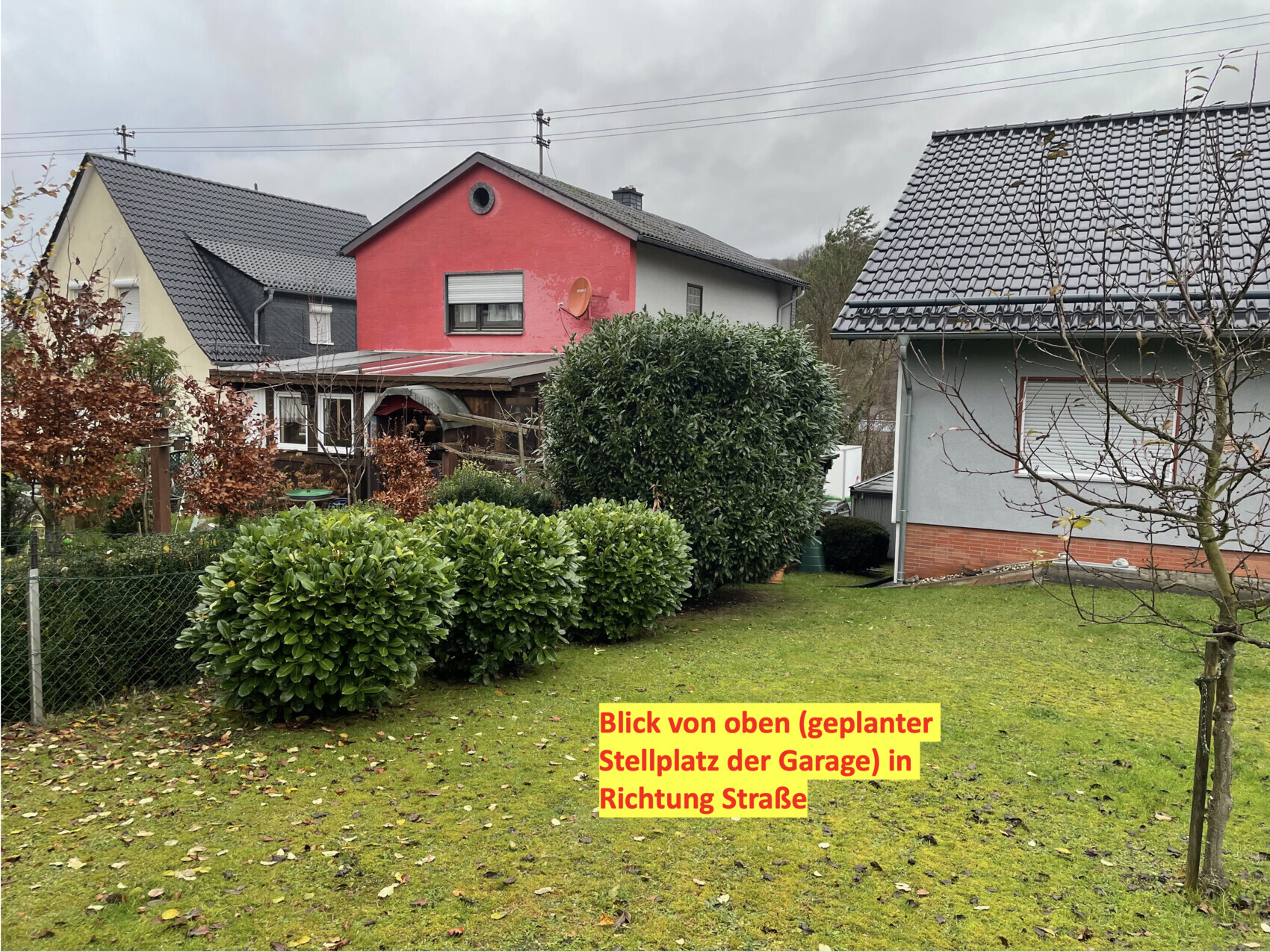 Vorgarten mit runden Hecken, drei Wohnhäuser (rotes zentrales Haus, links gelb, rechts grau).