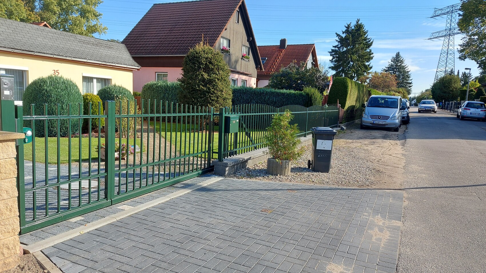 Grüner Metallzaun trennt gepflegten Vorgarten mit Hecken; geparkter Mercedes-Van am Straßenrand.
