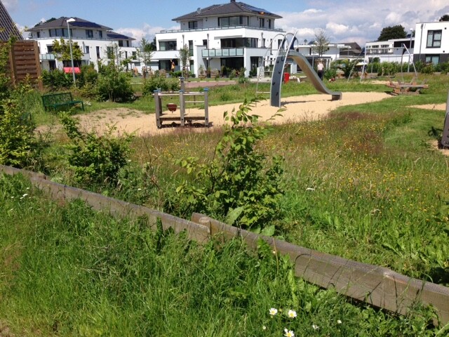 Moderner Spielplatz im Park mit Sandkasten und Spielgeräten; weiße Häuser im Hintergrund.