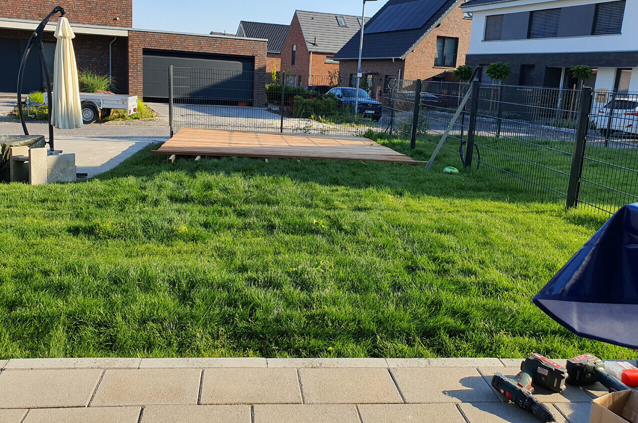 Garten mit grünem Rasen, Holzplattform auf Gestell, Zaun und Häuser im Hintergrund.