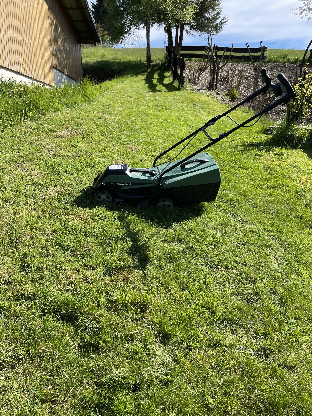 Grüner Rasenmäher im sonnigen Garten neben einem Holzhaus und Zaun.