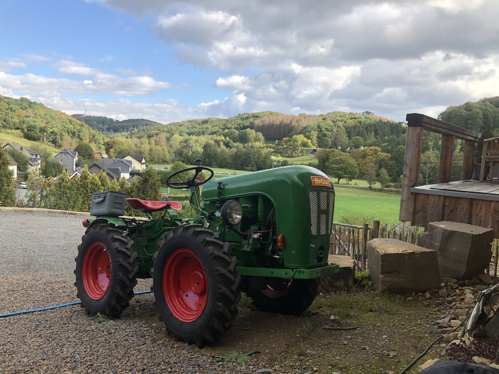 Grüner Vintage-Traktor steht auf Kies, vor ländlicher Hügellandschaft und Holzdeck.
