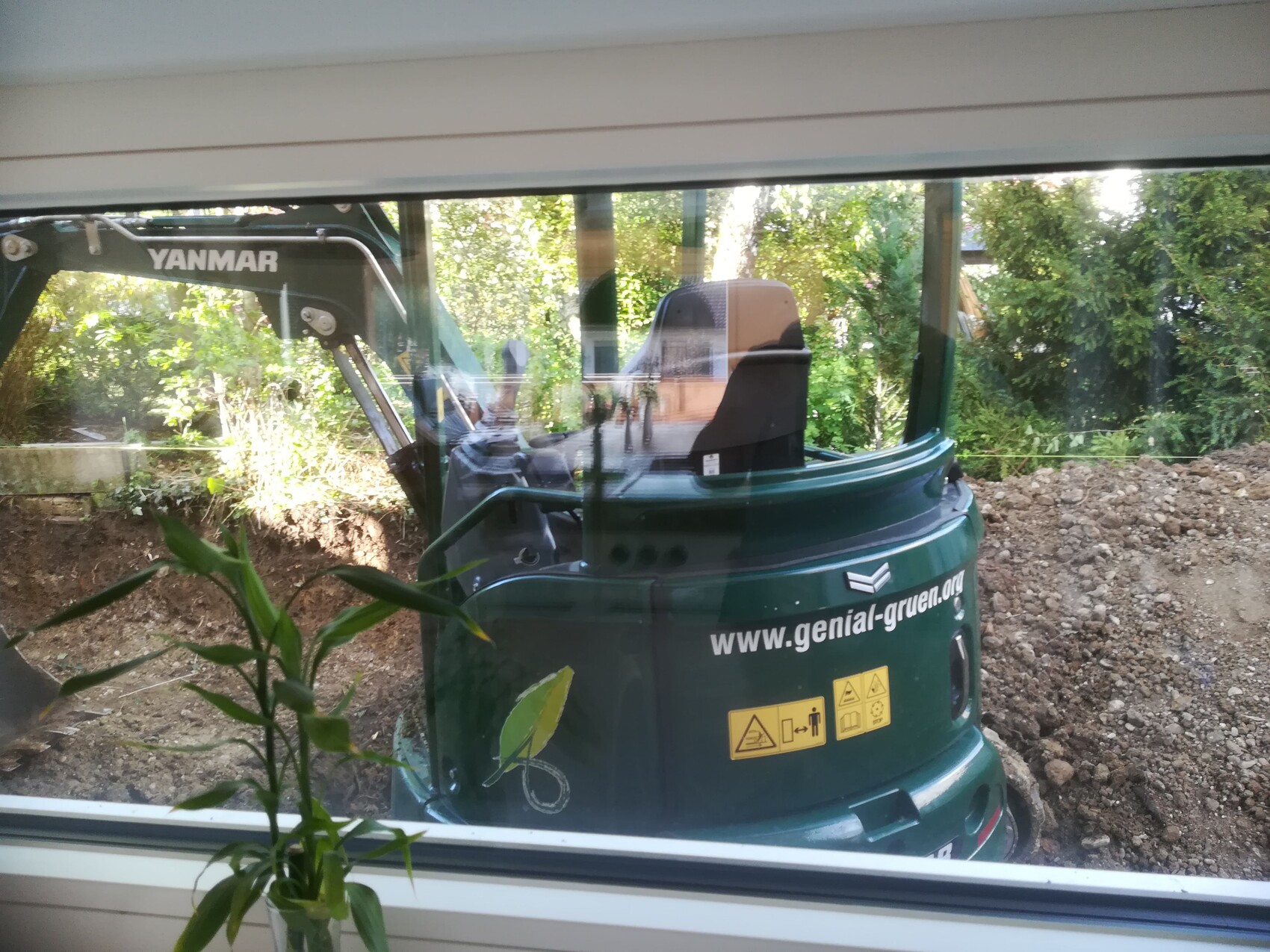 Grüne Yanmar-Baumaschine (Kleinbagger) vor Baugrube hinter Fensterscheibe sichtbar
