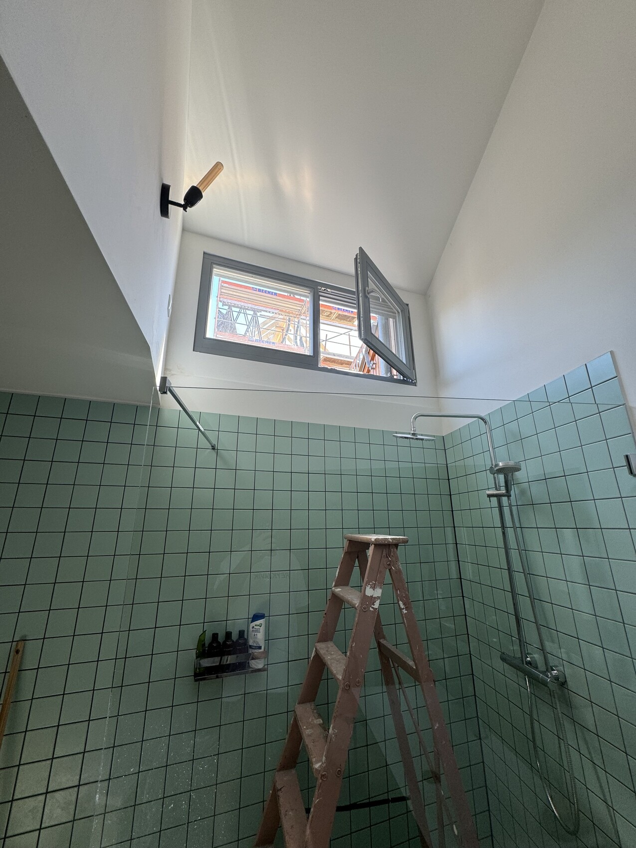 Badezimmer unter Renovierung mit grünen Fliesen, offenes Fenster, Holzleiter und Duscharmatur.