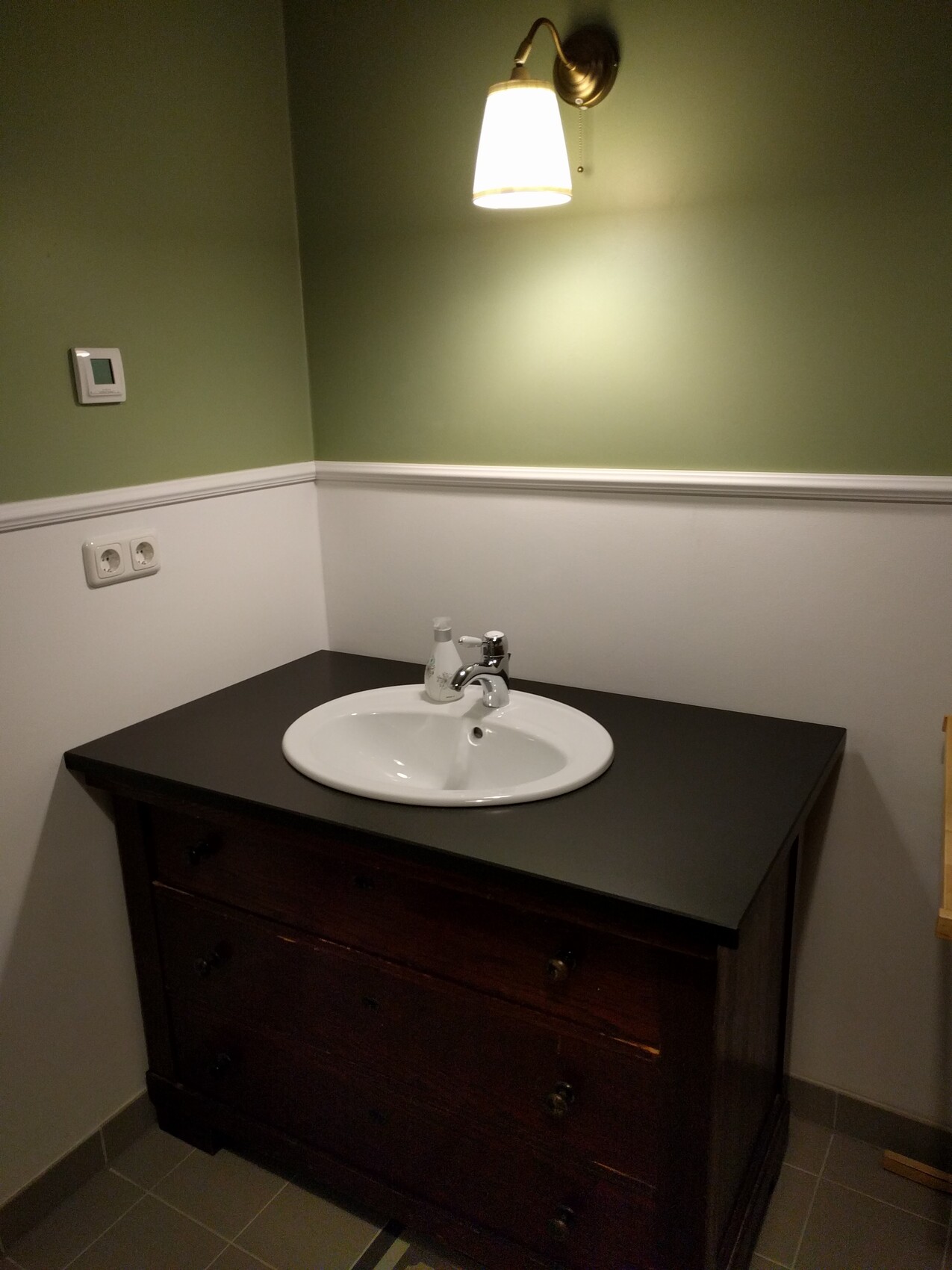Waschbecken aus weißer Keramik auf dunklem Holzunterbau, grüne Wand und Wandlampe darüber.