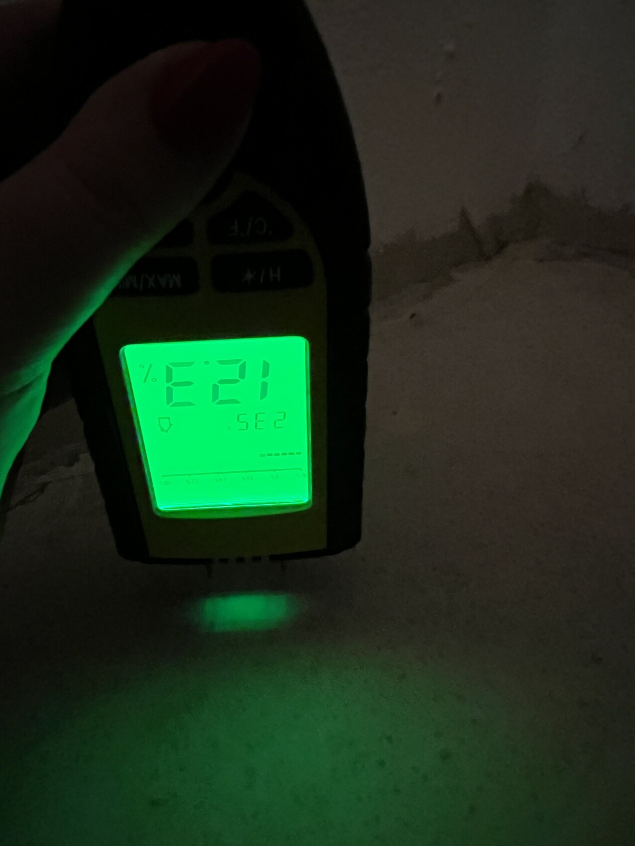 Hand hält ein digitales Messgerät mit grün beleuchtetem Display in dunkler Umgebung.