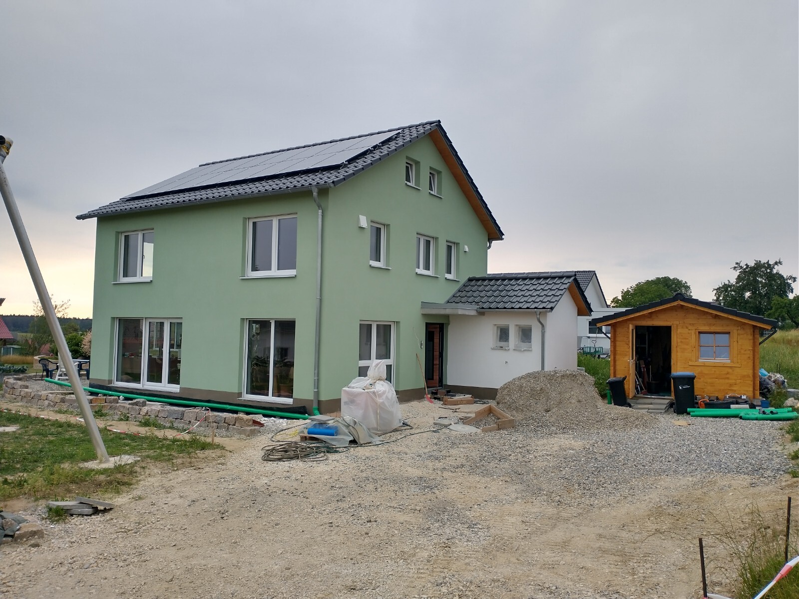 Zweistöckiges grünes Haus mit Solarzellen, Anbau und Holzschuppen auf Baustelle.