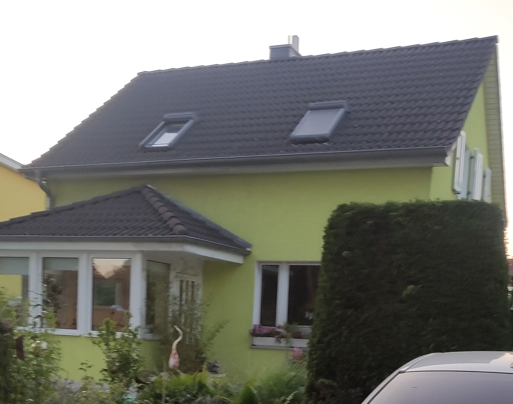 Grünes Einfamilienhaus mit dunklem Ziegeldach, Dachfenster, Garten und geparktem Auto.