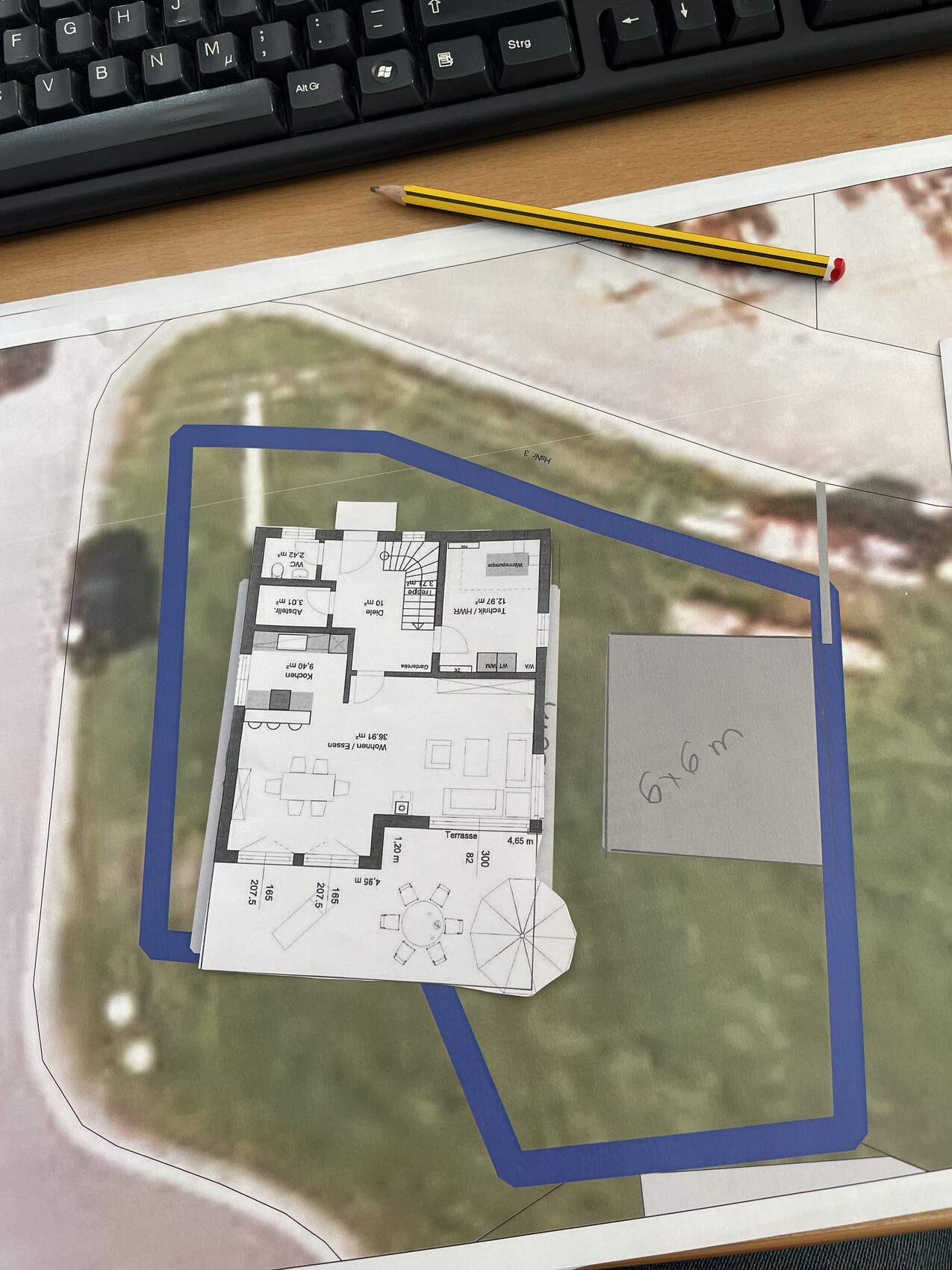 Papiermodell eines Hausgrundrisses auf Grundstückskarte mit Terrasse; daneben Bleistift.