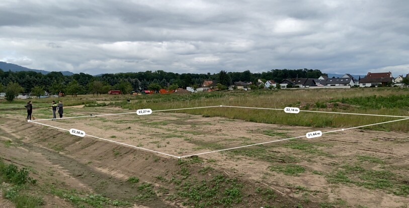 Geplantes Baugrundstück mit Vermessungslinien; zwei Personen am Rand, Häuser im Hintergrund.