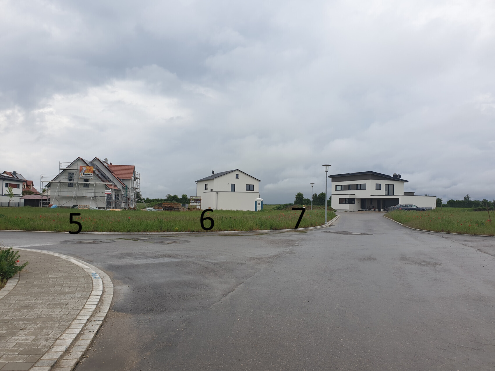 Baugrundstück mit eingerüstetem Haus links und modernem Haus rechts unter wolkigem Himmel