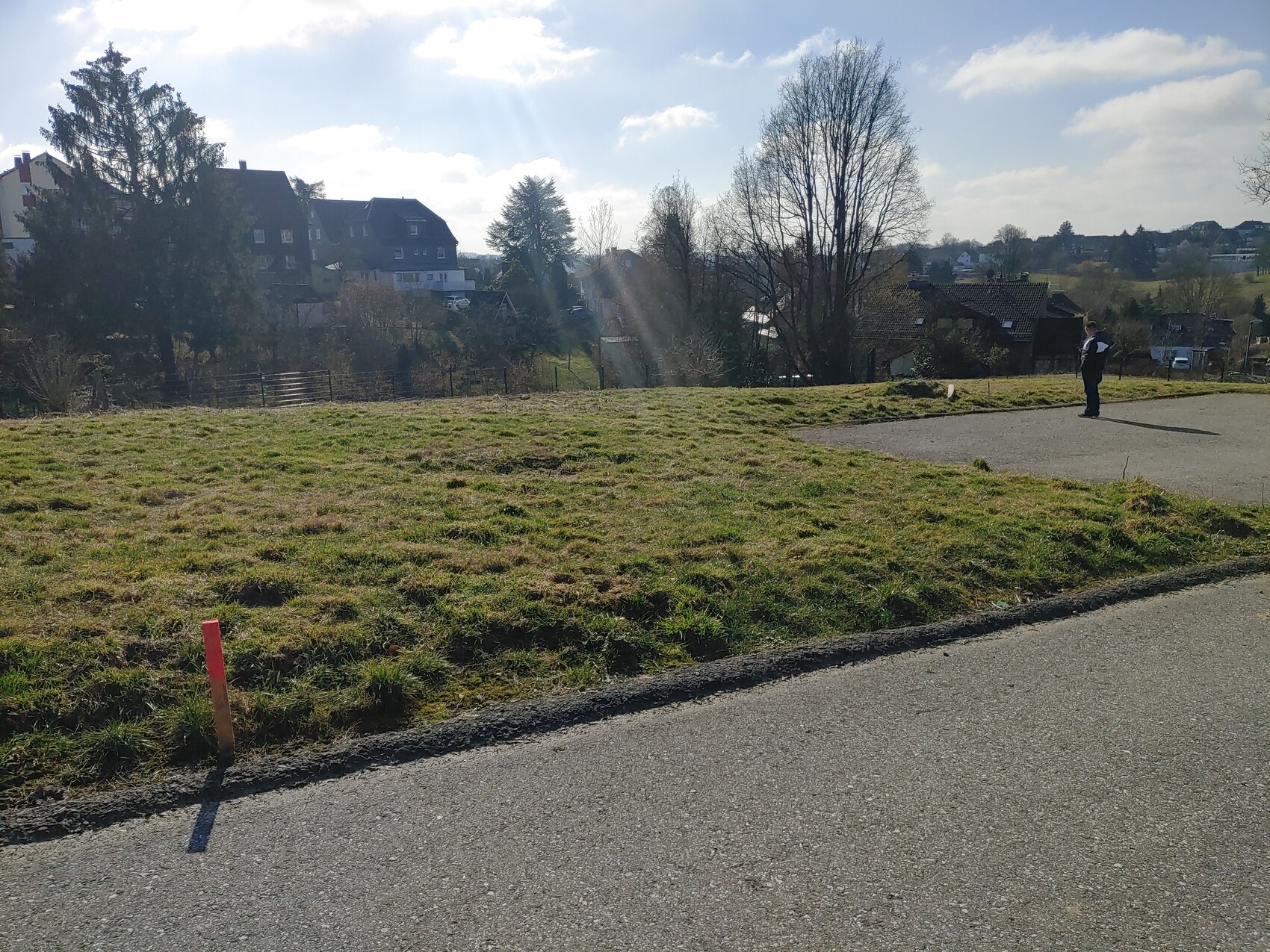 Mann steht auf einem gepflasterten Weg am grasigen Hang; roter Pfosten im Vordergrund, Häuser