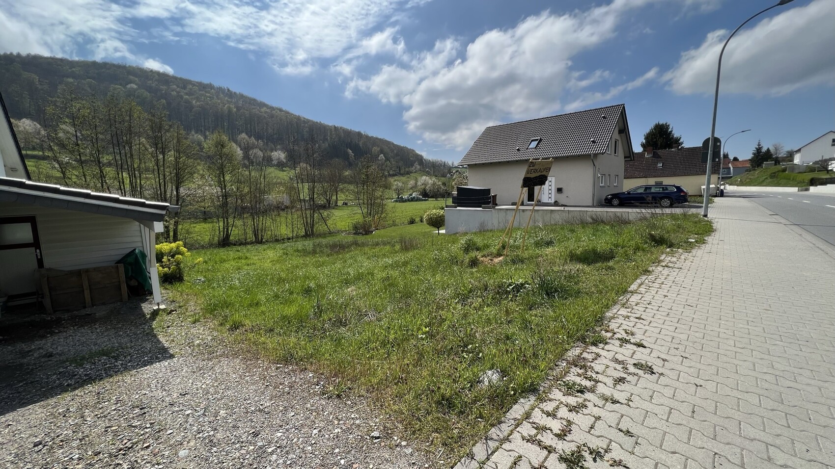 Wohnhaus am Straßenrand mit Schild 'Zu verkaufen' vor grasiger Wiese und Hügeln im Hintergrund.