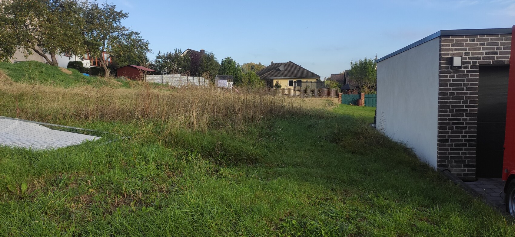 Grasiges, unbebautes Gelände mit Einfamilienhäusern im Hintergrund; rechts Ziegelwand.