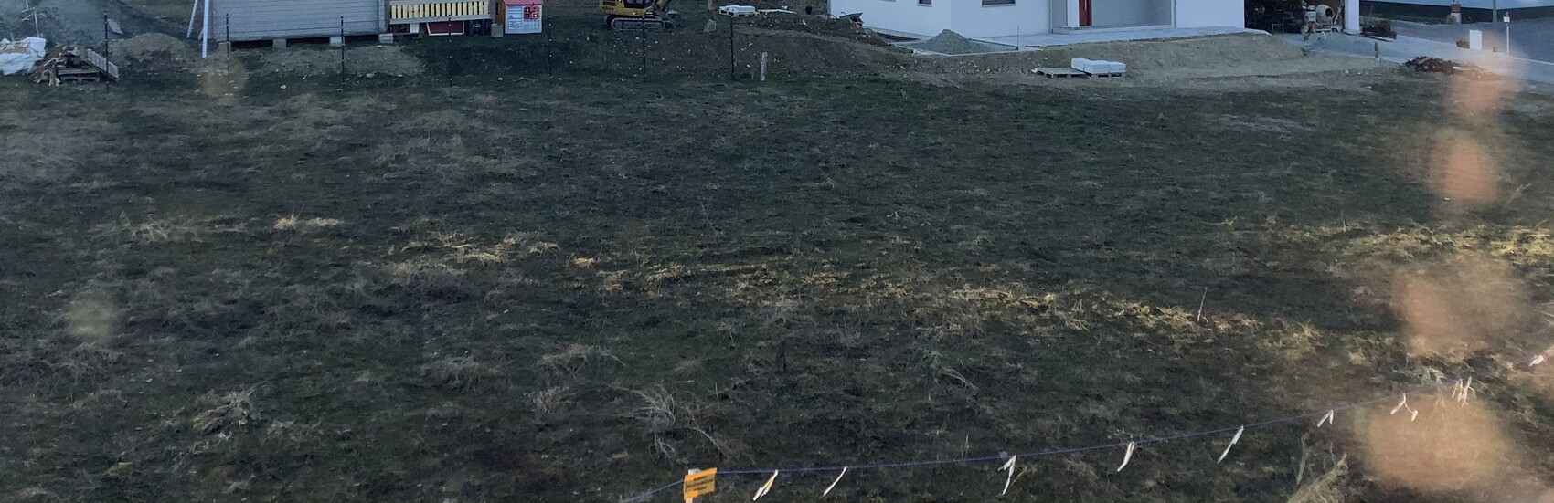 Weites Grasfeld mit Baugeräten, Zaunlinie und Gebäuden im Hintergrund.