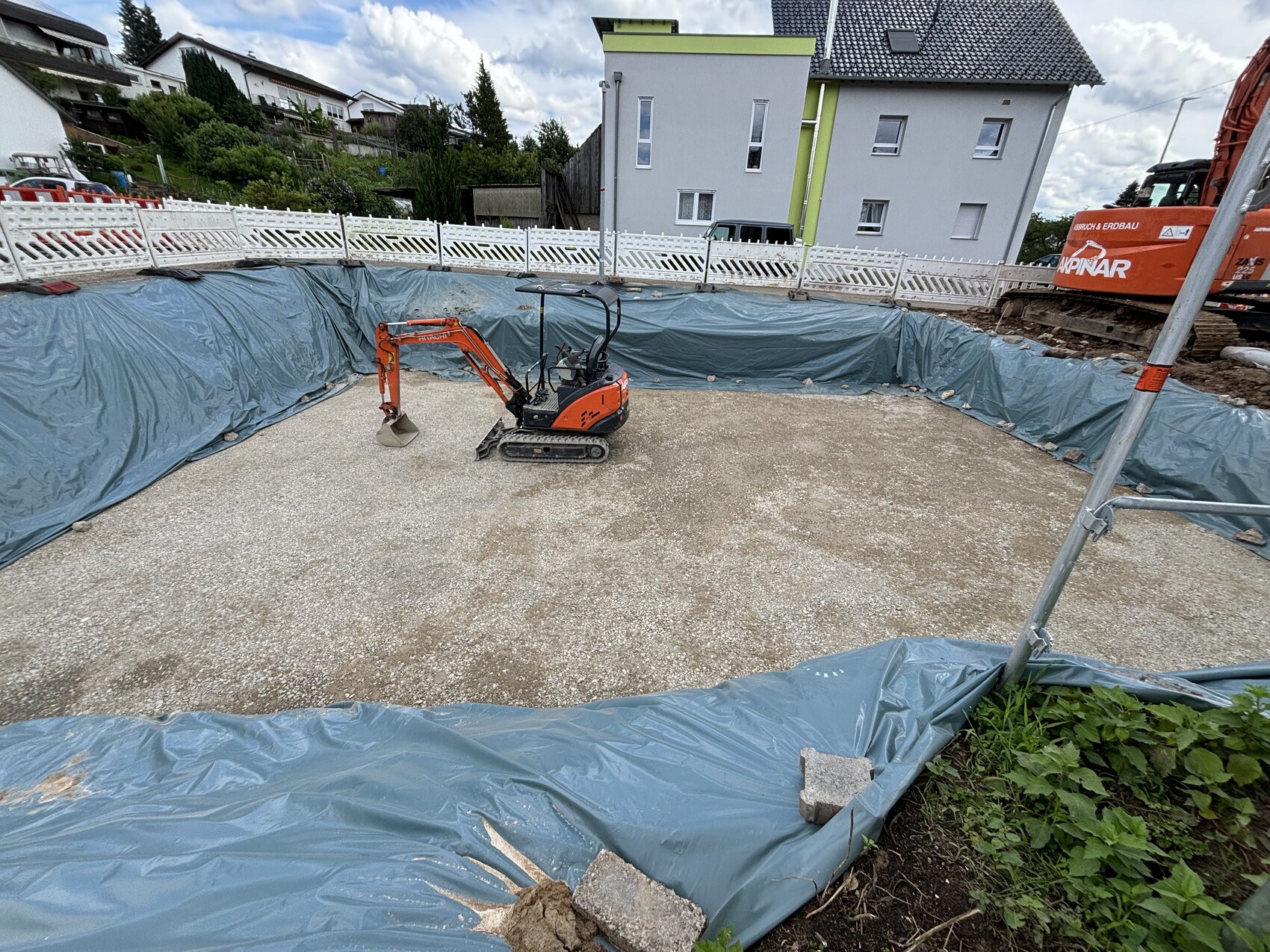 Baugrube mit orangefarbenem Mini-Bagger, U-förmig mit Planen ausgekleidet, Wohnhäuser im Hintergrund