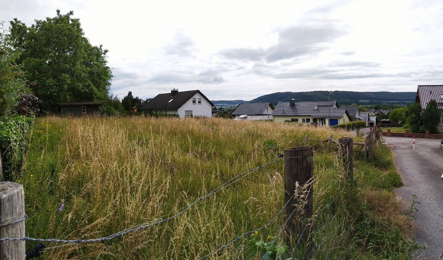 Weites Grasfeld am Straßenrand, Zaunpfosten mit Draht, Häuser und Hügel im Hintergrund.
