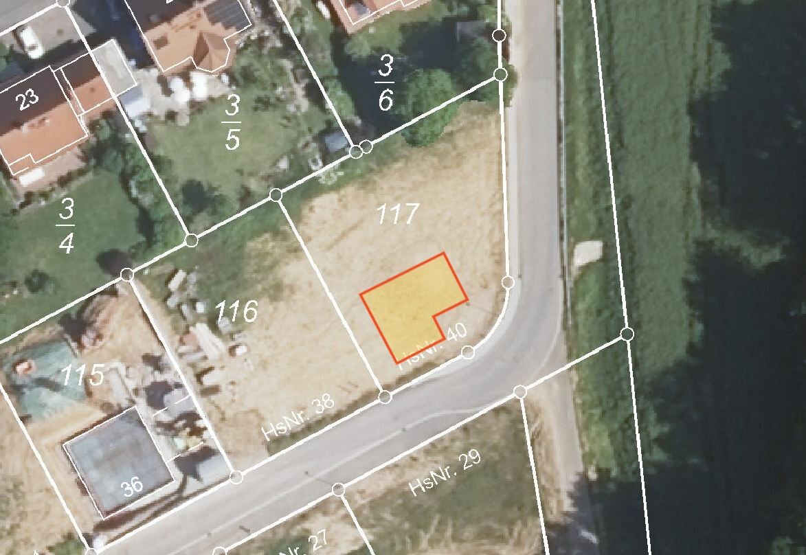 Luftbild eines Baugrundstücks mit rotem Umriss eines gelben Hauses neben kurviger Straße am Fluss.