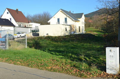 Freies Baugrundstück neben Wohnhäusern, grüne Wiese, Bauzaun und Infostele.