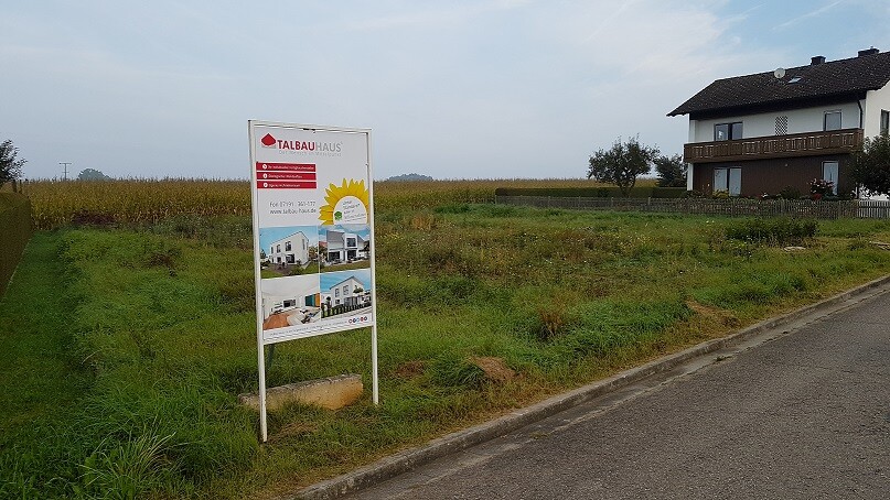 Baugrundstück mit Werbeschild am Straßenrand, angrenzendes Einfamilienhaus