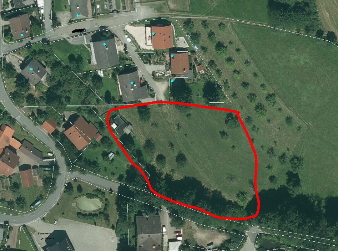 Luftbild einer grünen Parzelle rechts von Wohnhäusern; roter Umriss markiert das Gelände.