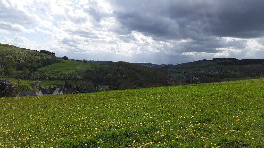 Grüne Wiesenlandschaft mit kleinem Dorf im Tal und wolkigem Himmel