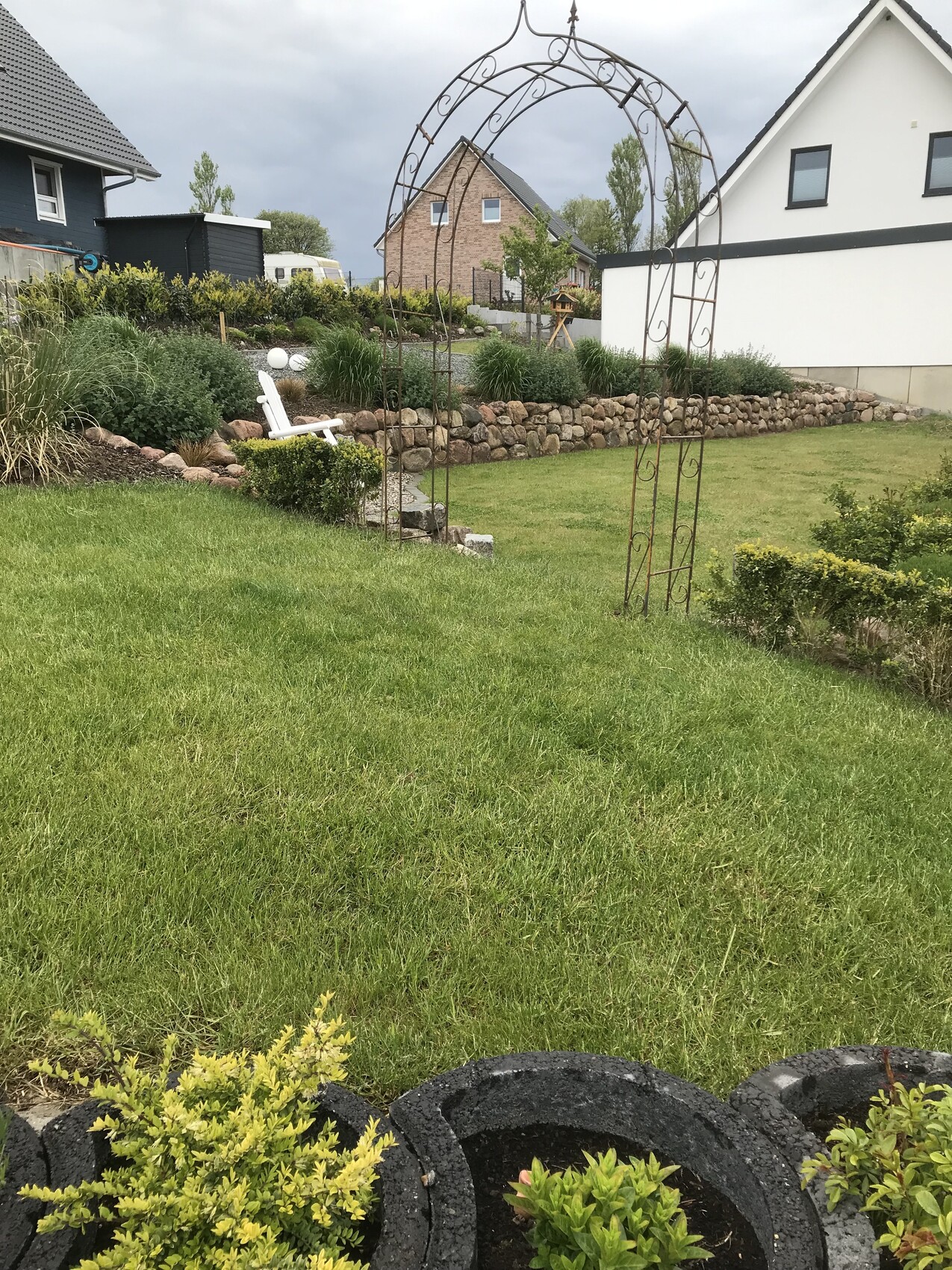 Garten mit schmiedeeisernem Torbogen, weißem Gartenstuhl und Steinmauer im Hintergrund
