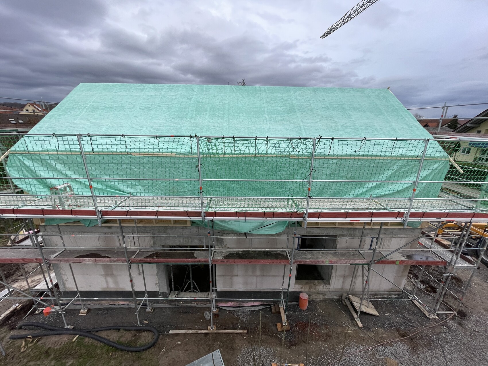 Baustelle mit Gerüst um ein Haus; grüne Abdeckplane über dem Dach, wolkiger Himmel.
