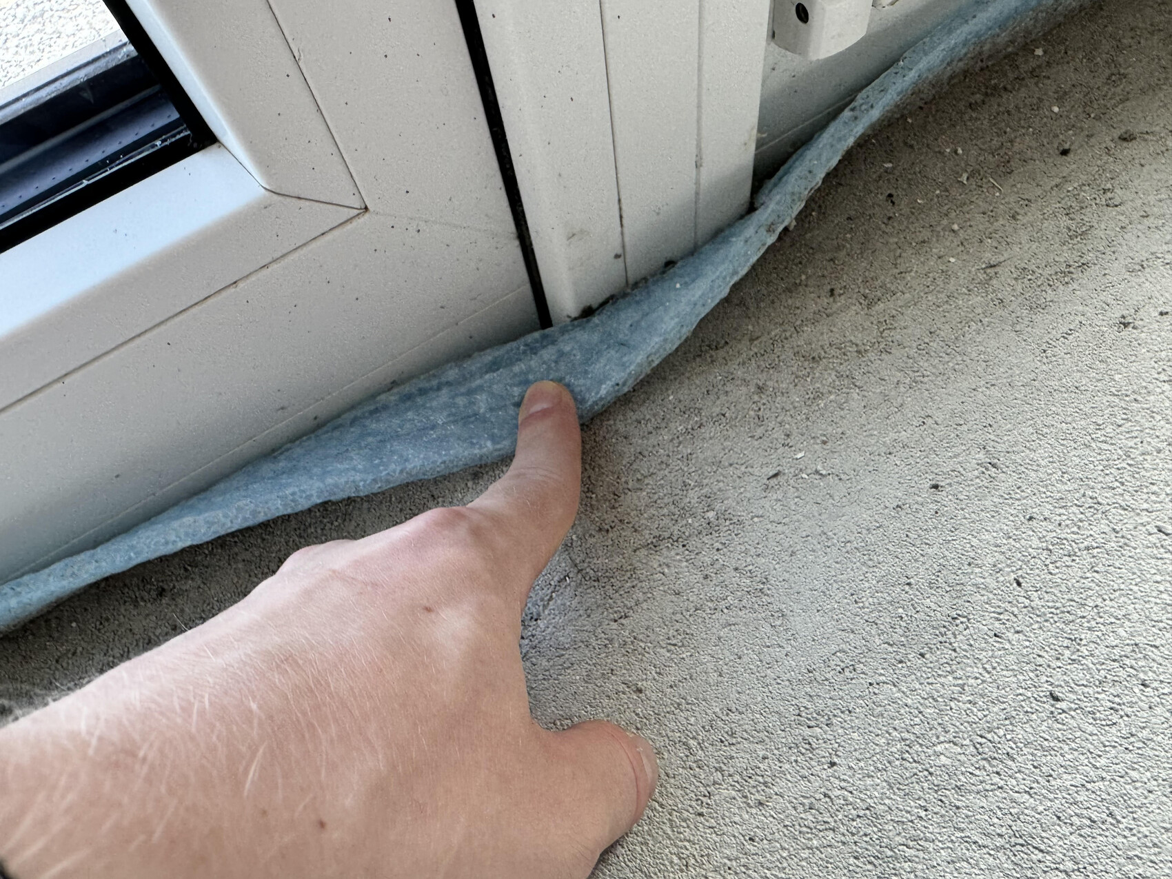 Ein Finger berührt eine blaue Dämmrolle am Fensterrahmen über rohem Betonboden.