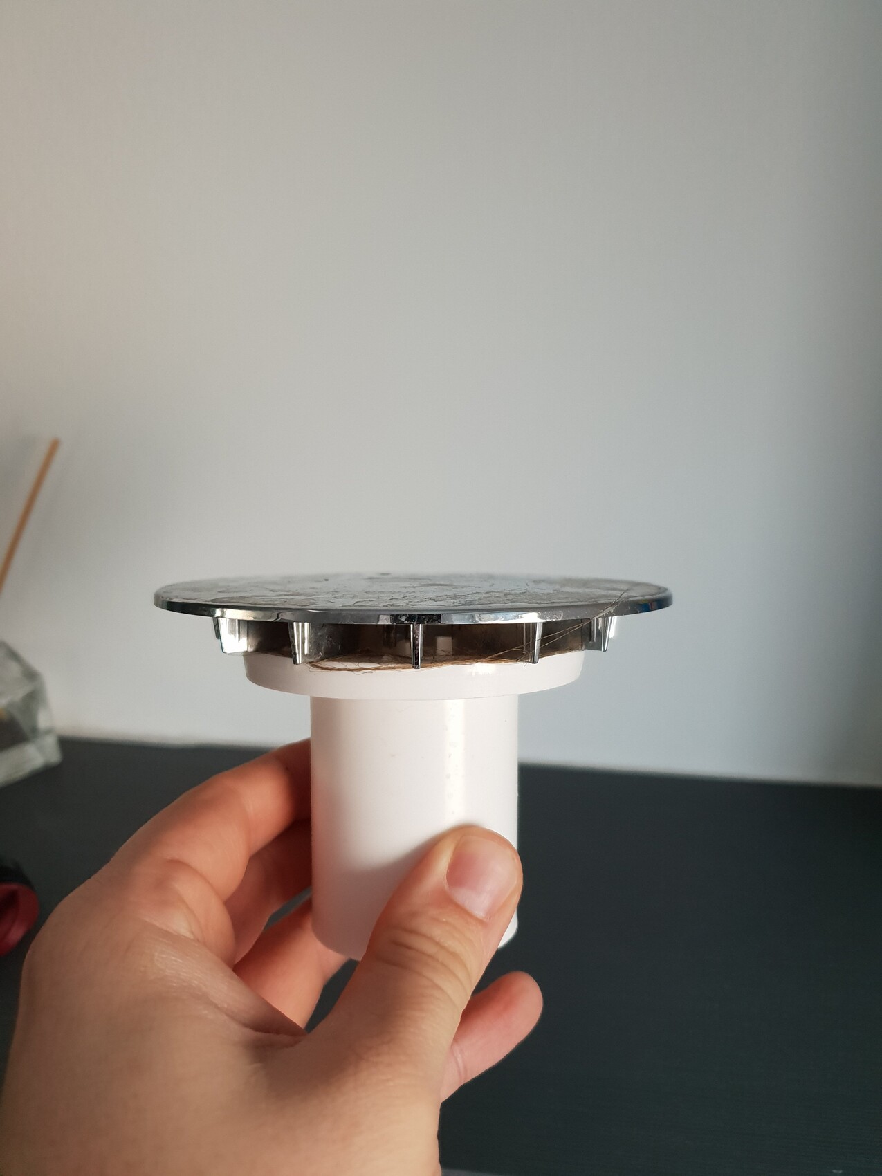 Hand hält weißen PVC-Abfluss mit runder Metallabdeckung vor neutraler Wand