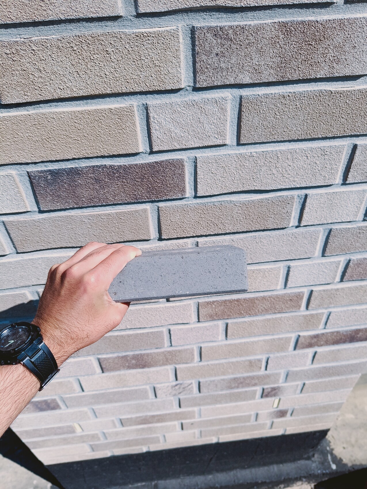 Eine Hand mit Armbanduhr hält einen grauen Ziegelblock gegen eine graue Ziegelwand.