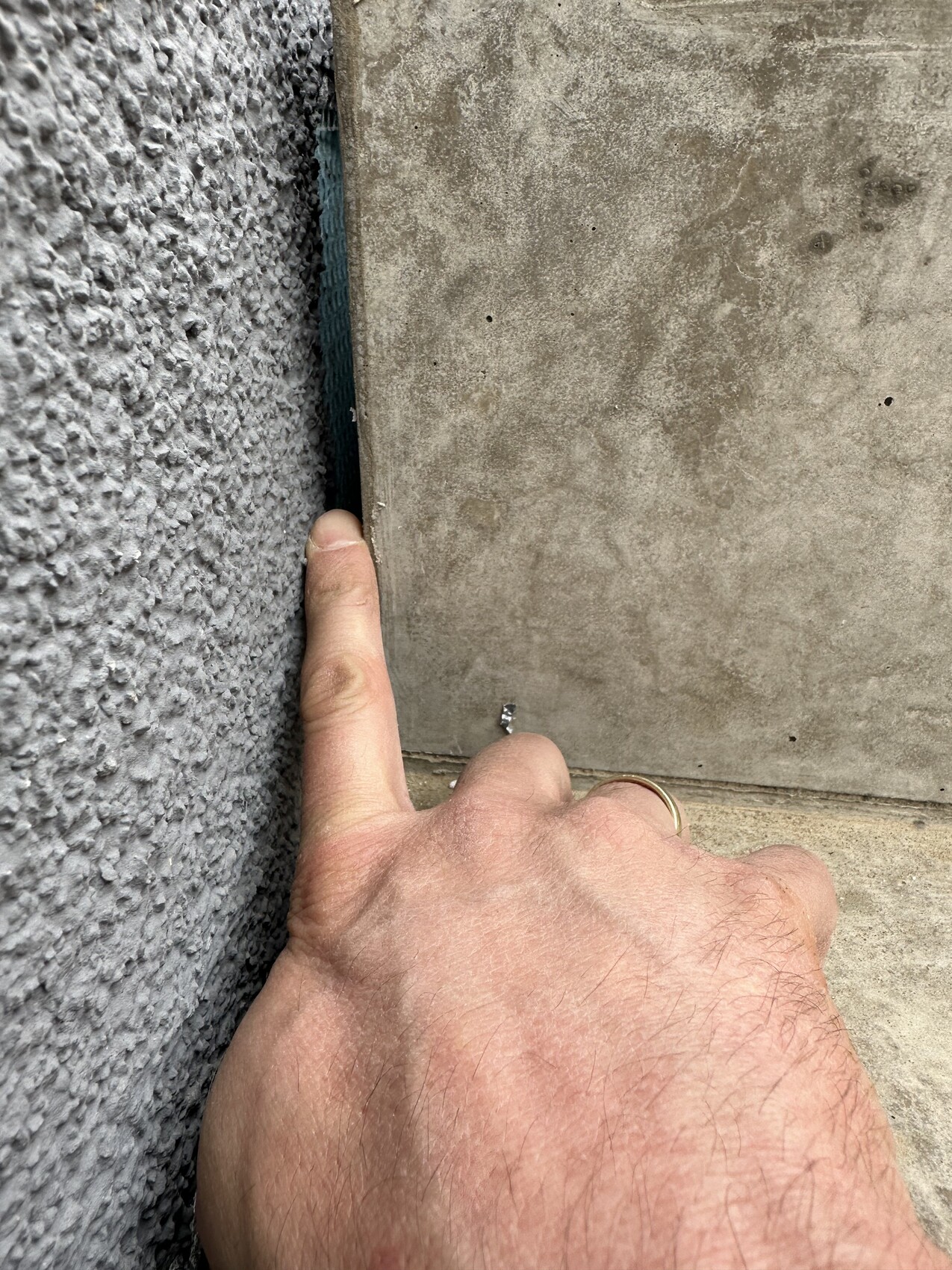 Hand mit Ring zeigt auf eine kleine Schraube im Spalt zwischen rauer Wand und Betonplatte.