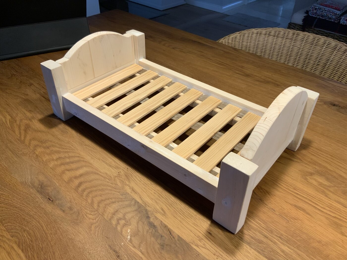 Kleines Puppenbett aus hellem Holz mit Lattenrost auf Holztisch.