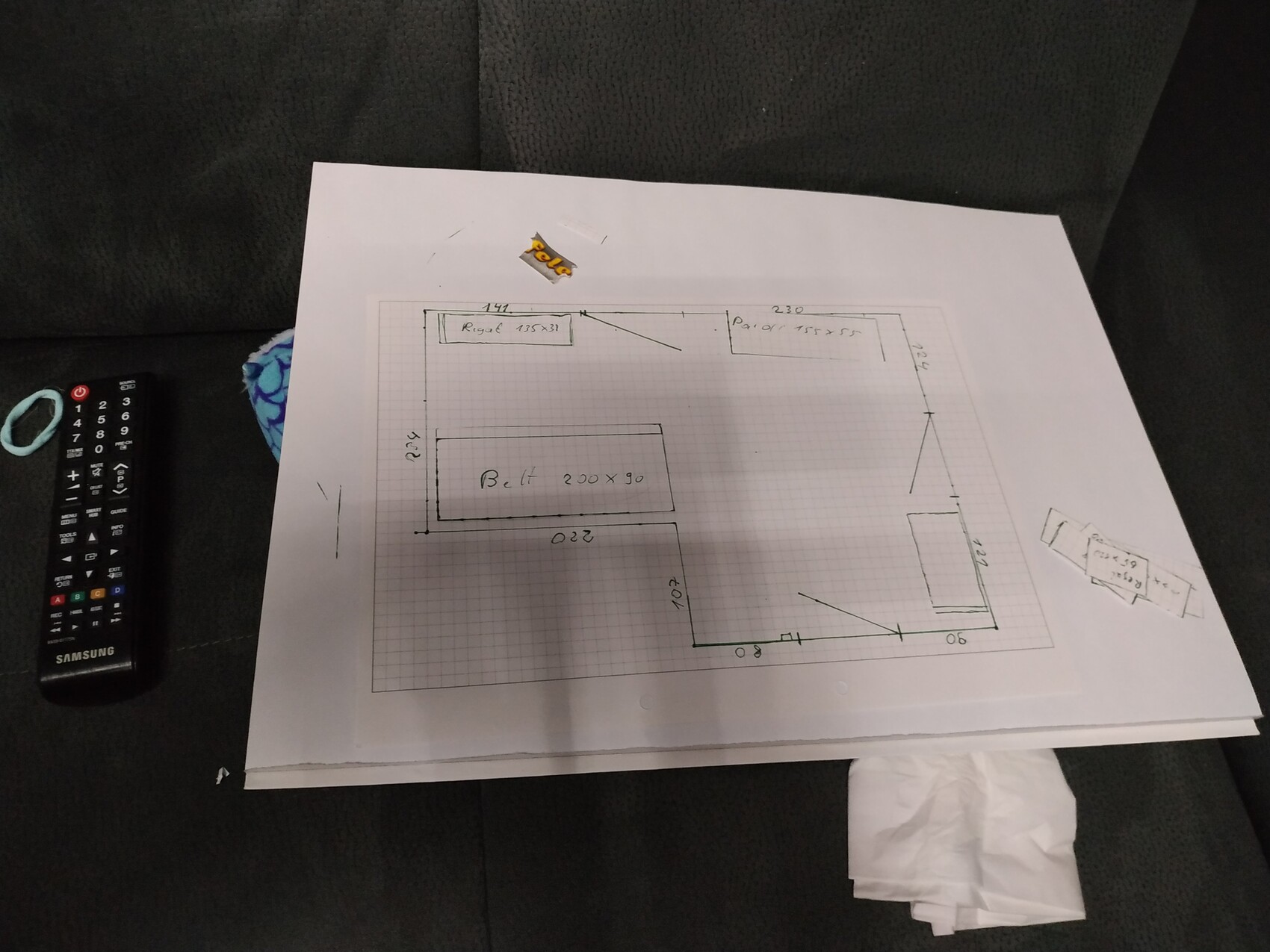 Handgezeichnete Grundrissskizze mit Raumaufteilung auf Papier, neben Fernbedienung auf dunklem Sofa