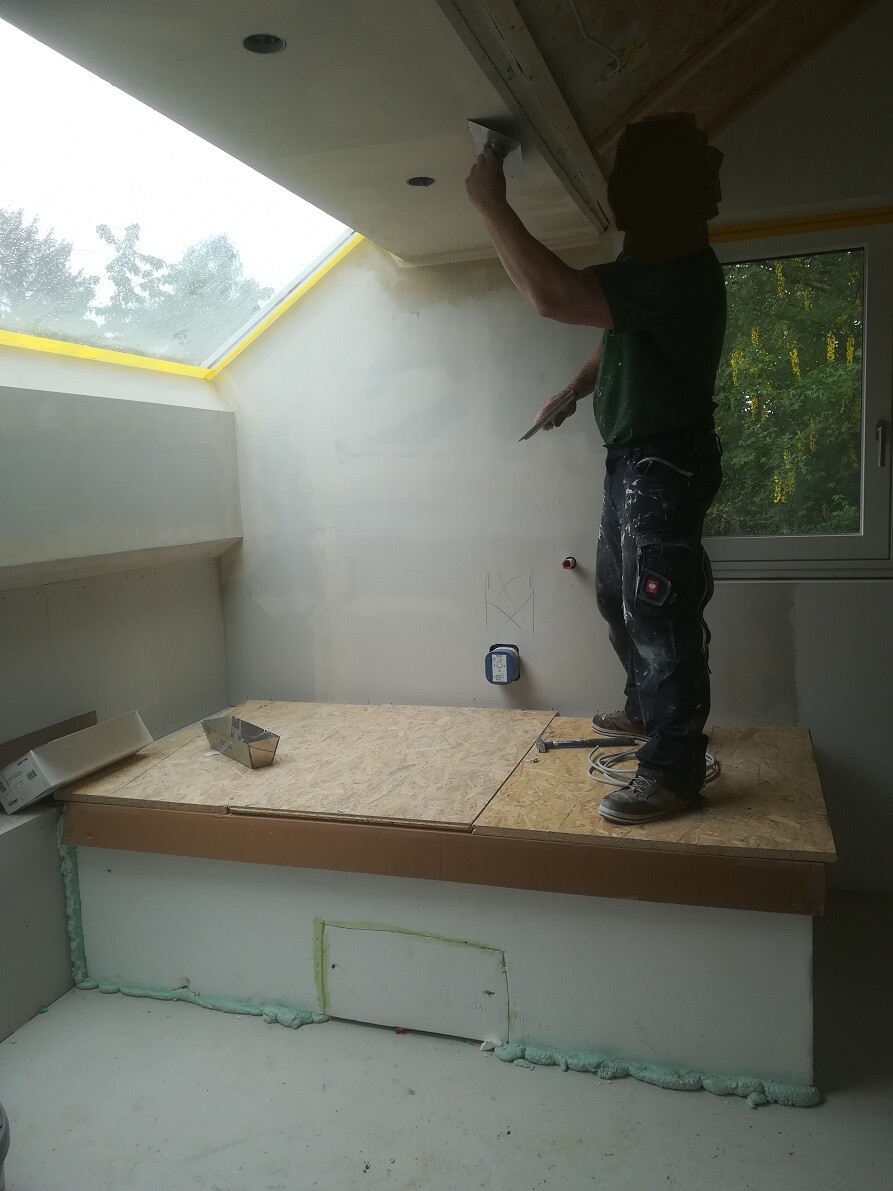 Mann verputzt die Decke, steht auf einer Holzplattform in einem Renovierungsraum.
