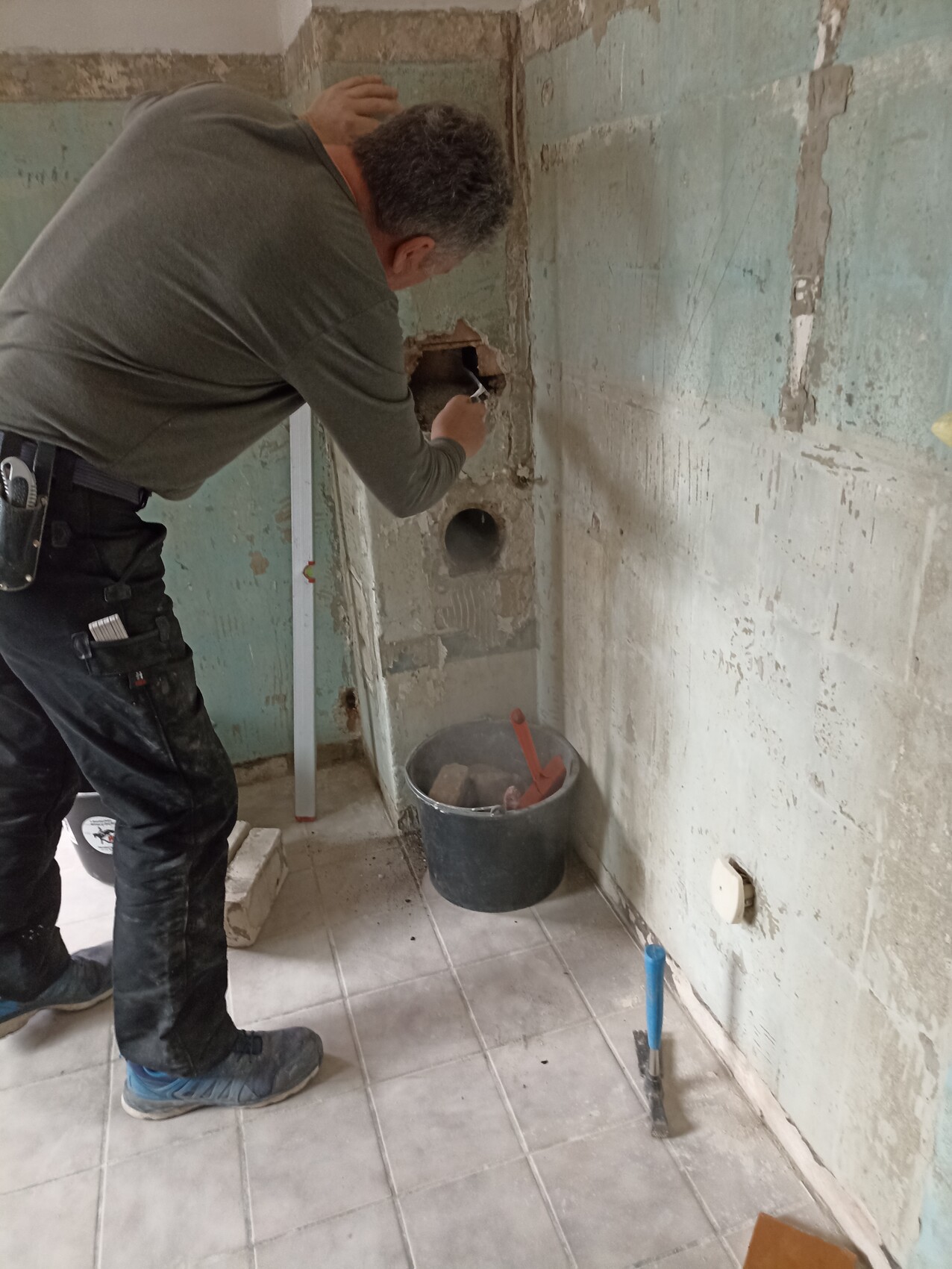 Handwerker entfernt Putz aus Wand mit Meißel; Eimer, Level und Fliesenboden sichtbar.
