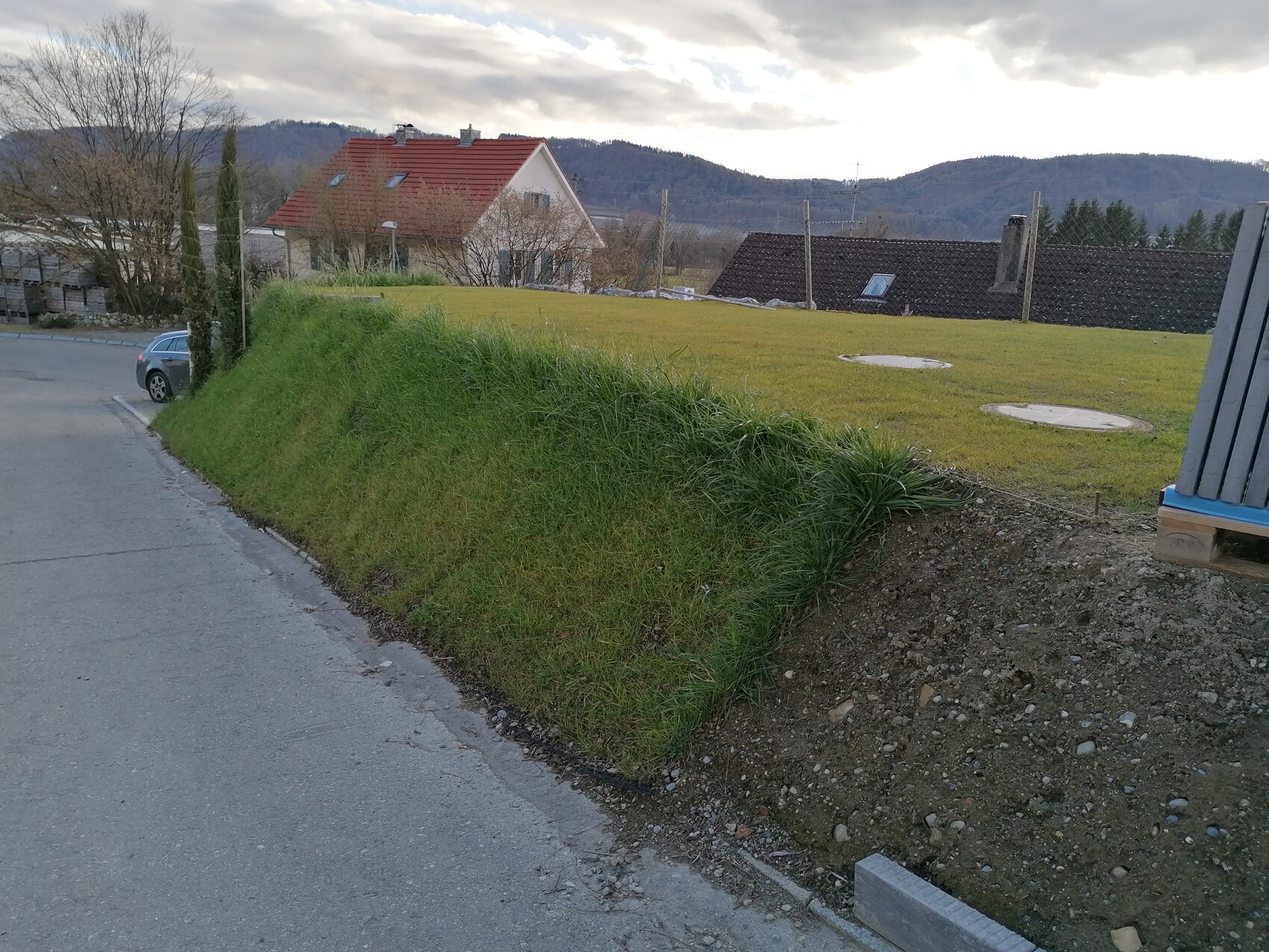 Grasbewachsener Erdwall neben Straße; Auto am Rand; Haus mit rotem Dach und Hügel im Hintergrund.