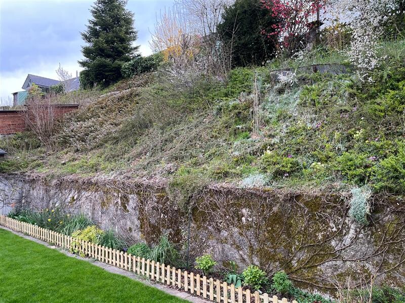 Garten mit steilem bepflanztem Hang über einer Steinmauer; am Fuß Holzzaun und Gras.