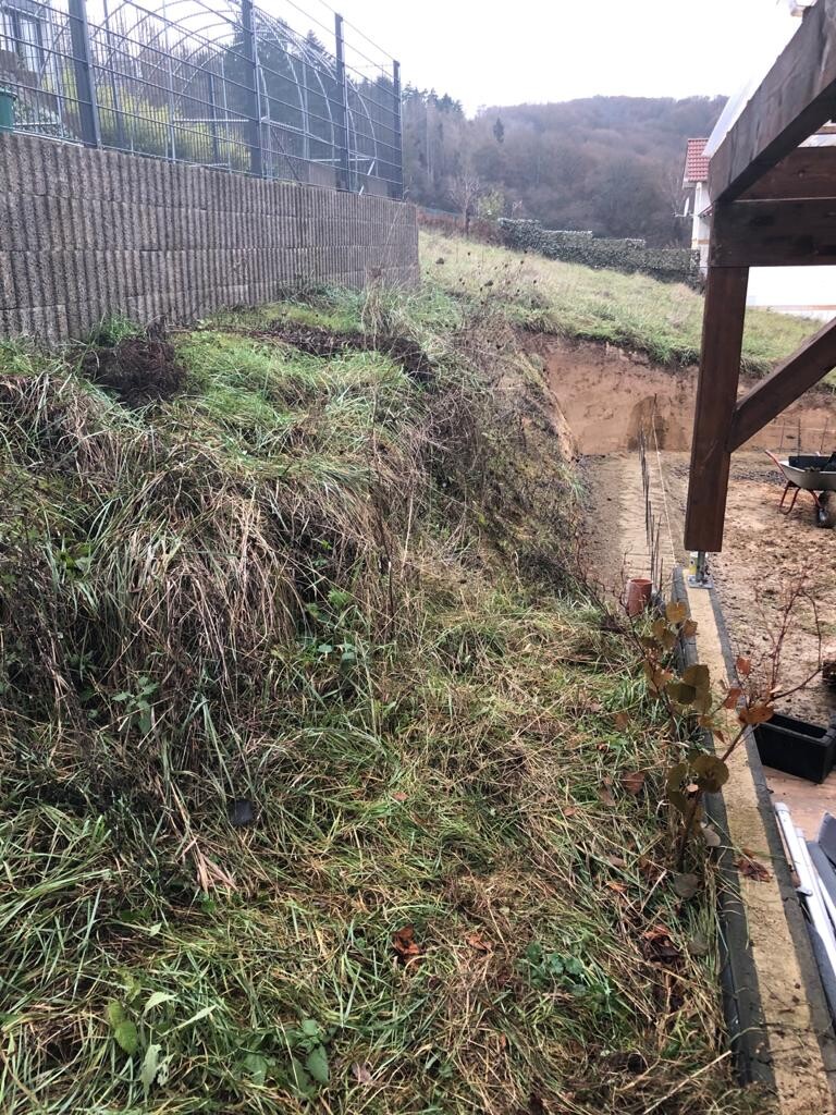 Grasbewachsene Böschung am Hang neben einer Betonmauer mit Geländer; rechts Bauholz und Werkzeuge.
