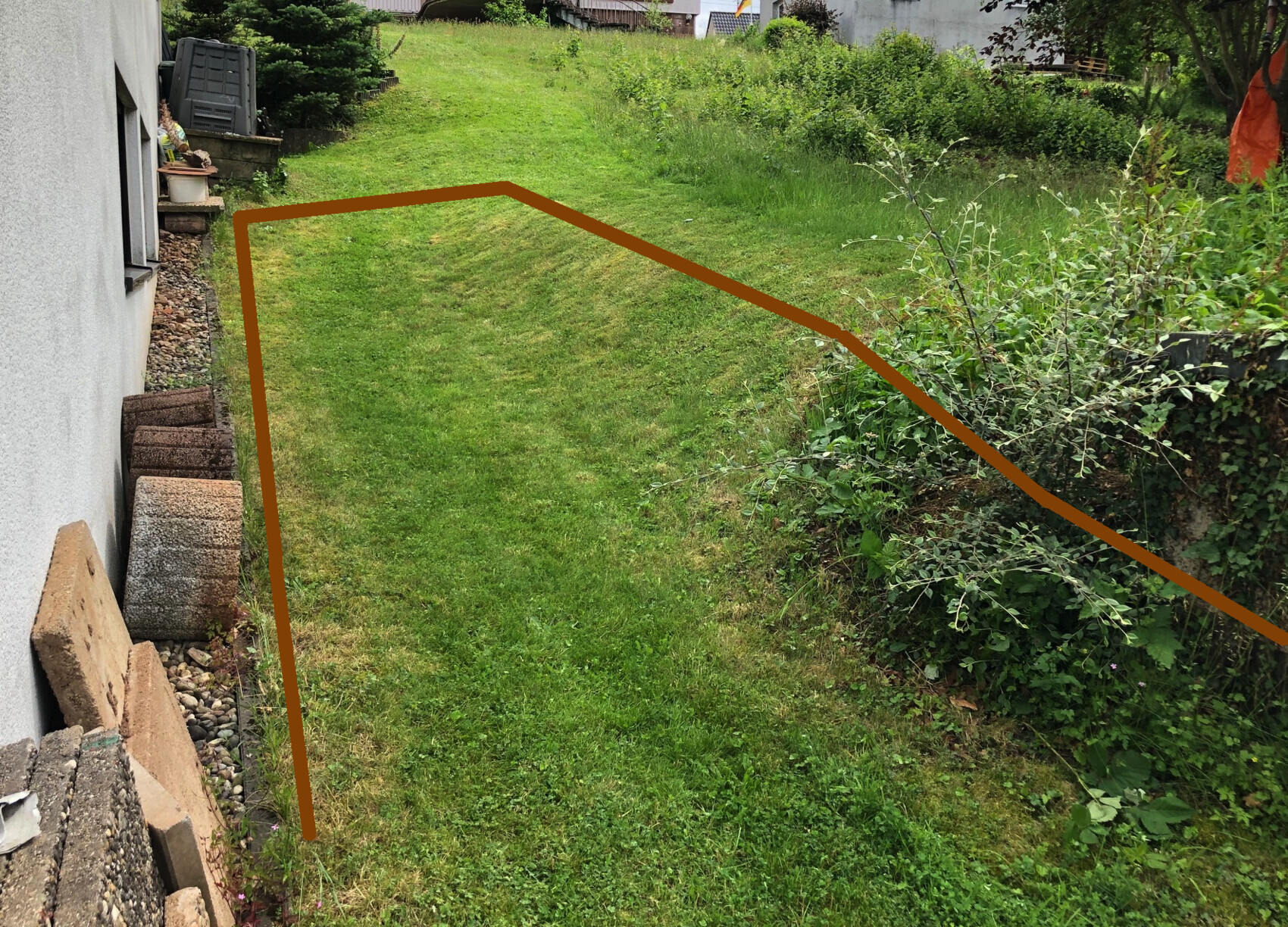 Garten mit leicht geneigtem Rasen, links Hauswand und Steine, braune Markierung einer Fläche
