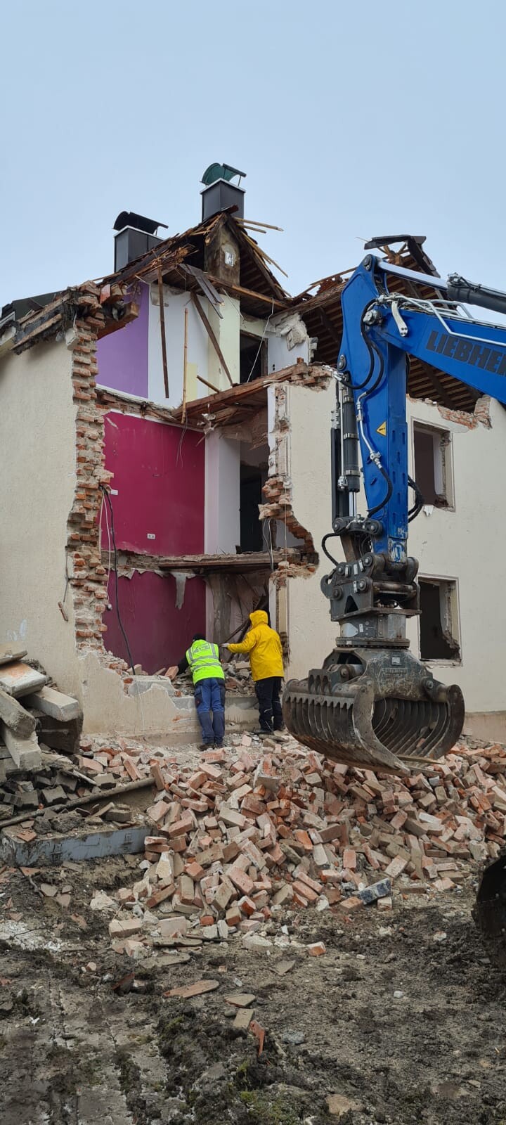 Baustelle: Hausabriss mit Kranarm, zwei Arbeiter in Neonjacken, Ziegeltrümmer am Boden