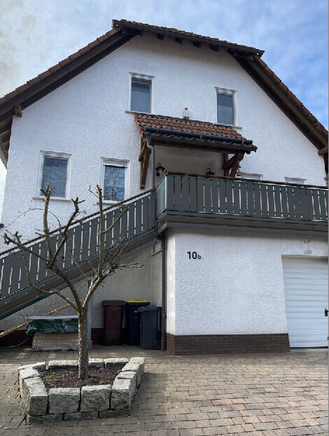 Zweistöckiges weißes Haus mit Balkon, Außentreppe, Baum im Steinbeet und Garage.
